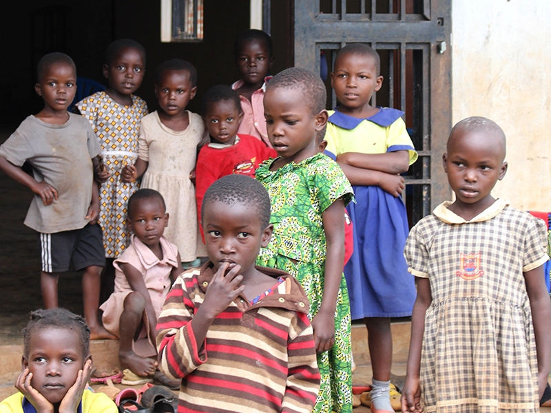 helfende hände - Eine Gruppe kleiner Kinder in unterschiedlich gemusterten Kleidern und Hemden steht und sitzt vor einem Gebäude; einige blicken in die Kamera, hinter ihnen sind eine Türöffnung und ein vergittertes Fenster zu sehen. helfende hände ggmbh hamburg