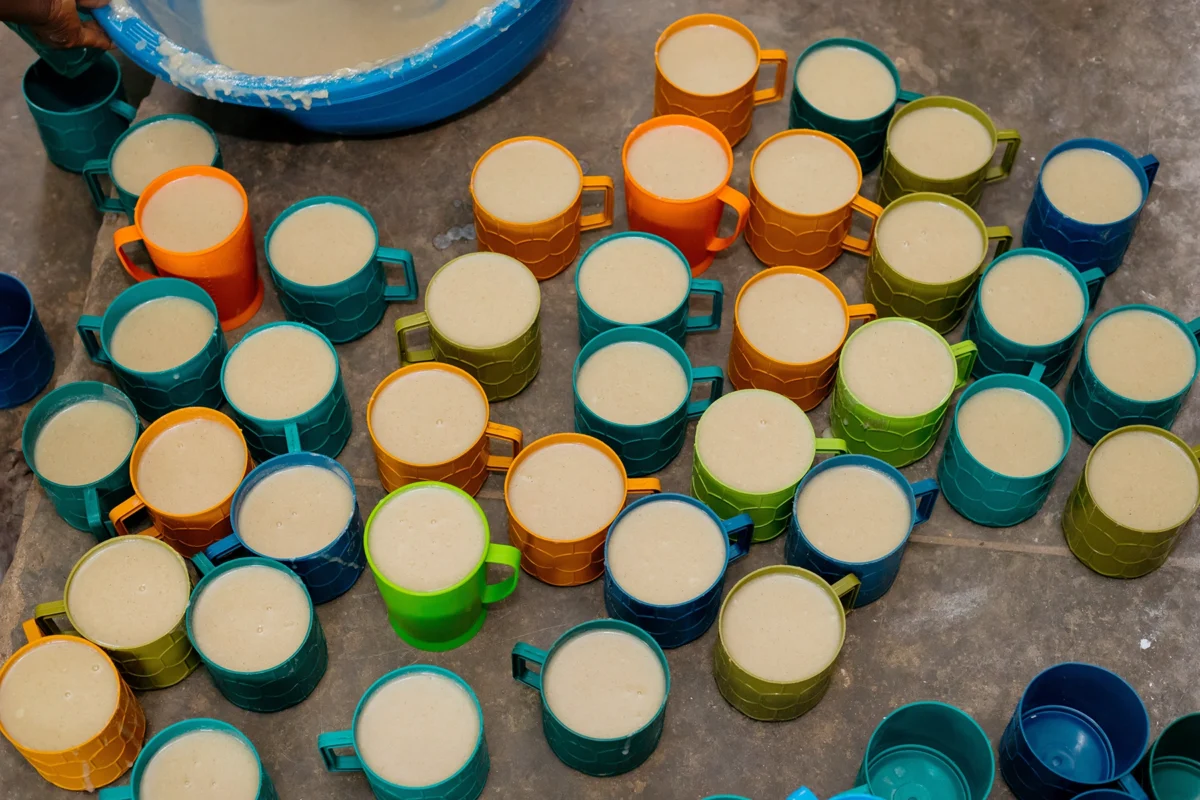 helfende hände - Auf einer Fläche sind mehrere Plastikbecher in den Farben Blau, Orange, Grün und Gelb angeordnet. Jeder Becher ist mit einer hellbeigen Flüssigkeit gefüllt, und in der Ecke ist teilweise eine große Schüssel mit einer ähnlichen Flüssigkeit zu sehen. helfende hände ggmbh hamburg