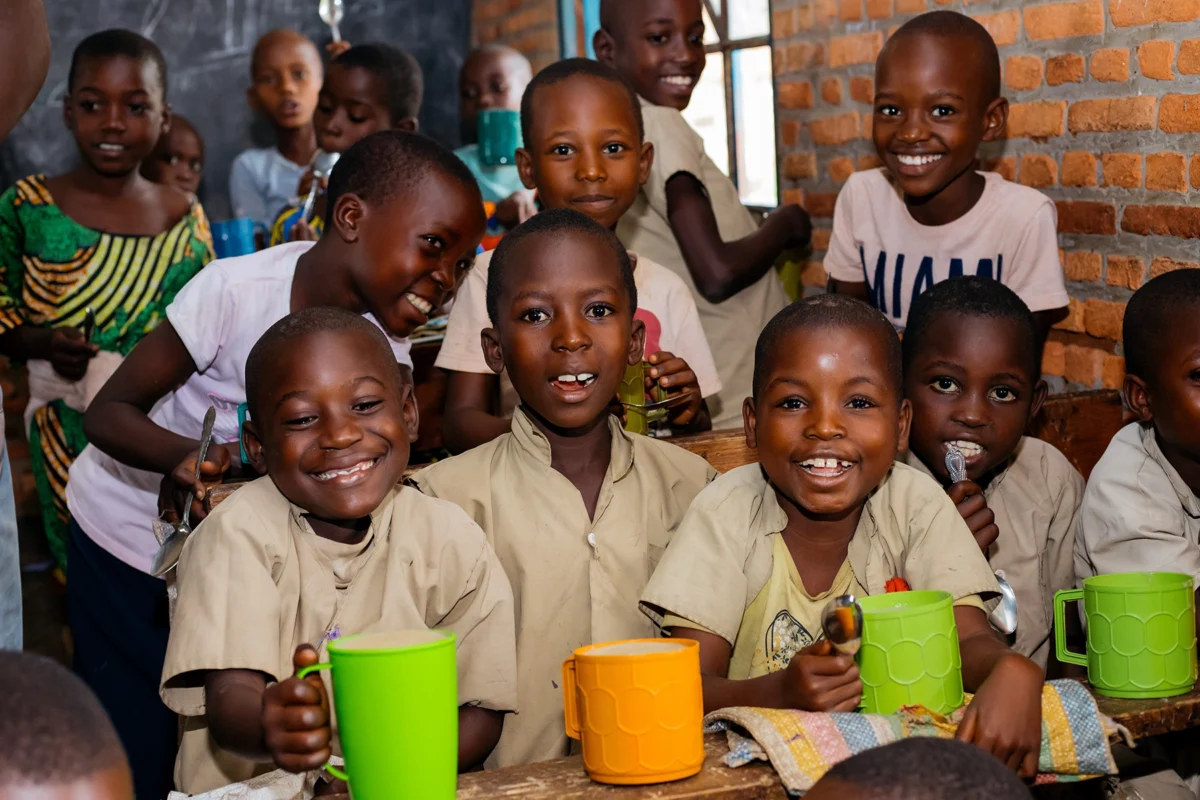 helfende hände - Eine Gruppe von Kindern sitzt dicht beieinander an den Schreibtischen in einem Klassenzimmer und lächelt in die Kamera. Sie halten bunte Becher und Löffel in den Händen. Im Hintergrund stehen weitere Kinder. Das Klassenzimmer hat Backsteinwände und eine Tafel. helfende hände ggmbh hamburg