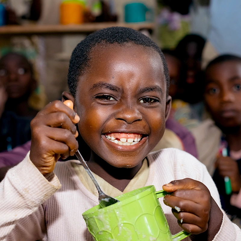 helfende hände - Ein lächelndes Kind hält einen grünen Becher und einen Löffel in der Hand; im unscharfen Hintergrund sind mehrere andere Kinder zu sehen. Das Kind trägt ein helles Hemd und scheint gerade eine Mahlzeit oder ein Getränk zu genießen. helfende hände ggmbh hamburg