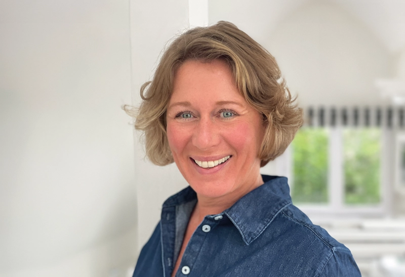helfende hände - Annette Freckmann, mit kurzen, welligen blonden Haaren, lächelt in die Kamera. Sie trägt ein blaues Jeanshemd und steht in einem hellen Raum mit weißen Wänden und großen Fenstern im Hintergrund. helfende hände ggmbh hamburg