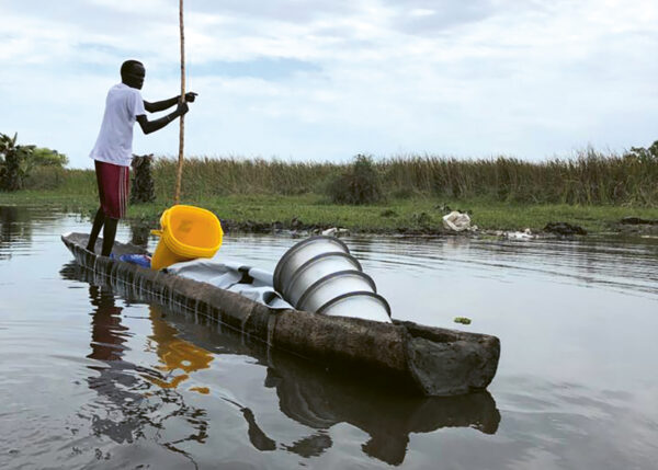 helfende hände - Eine Person steht auf einem hölzernen Kanu im seichten Wasser und steuert es mit einer langen Stange. Auf dem Kanu befinden sich mehrere Metallbehälter und gelbe Plastikeimer. Im Hintergrund sind hohes Gras und ein bewölkter Himmel zu sehen. helfende hände ggmbh hamburg