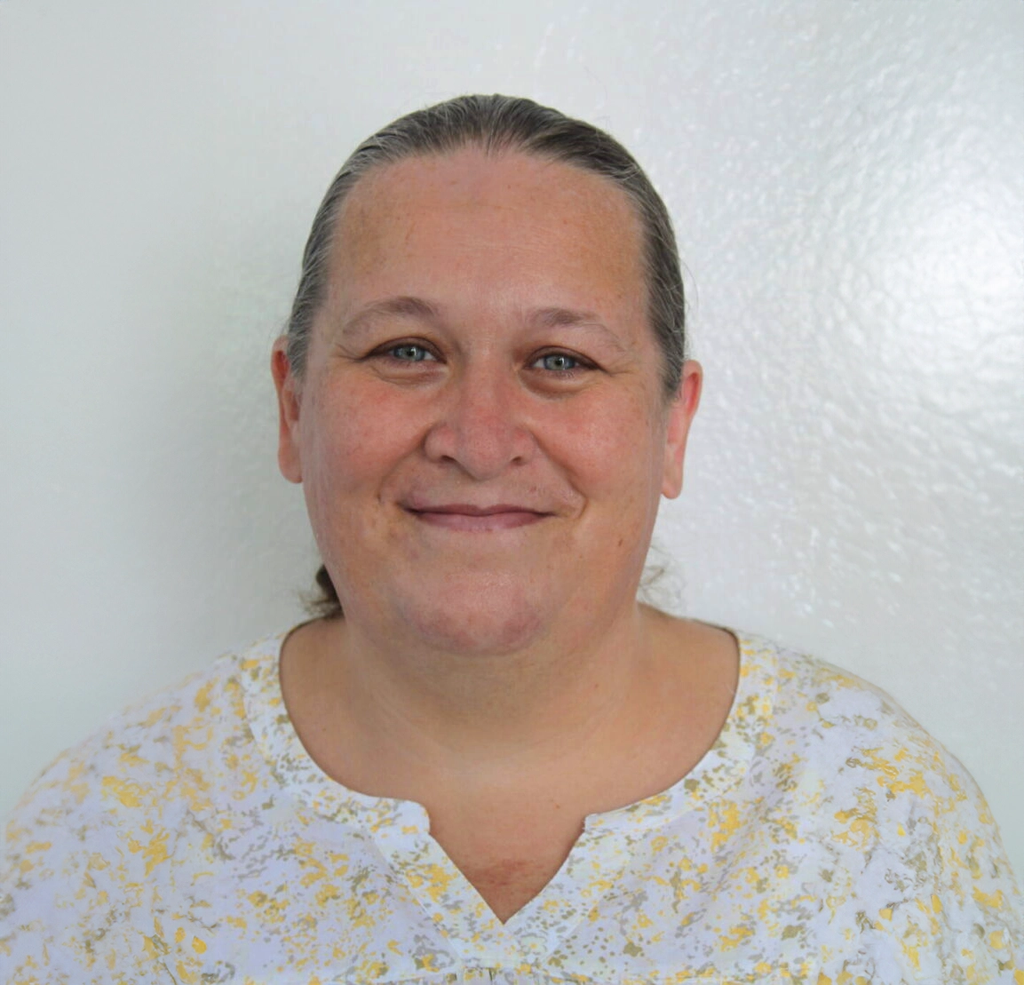 helfende hände - Licia Betor, mit heller Haut und nach hinten gebundenen braunen Haaren, lächelt in die Kamera. Sie trägt ein weißes Hemd mit gelbem Muster vor einem schlichten weißen Wandhintergrund. helfende hände ggmbh hamburg