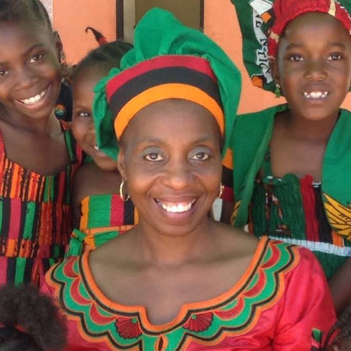 helfende hände - Shani Britton steht in einem farbenfrohen traditionellen Kleid und einem Kopftuch da und lächelt in die Kamera. Umgeben von Kindern in gleichförmigen, gemusterten Outfits posieren sie alle fröhlich vor einer orangefarbenen Wand. helfende hände ggmbh hamburg