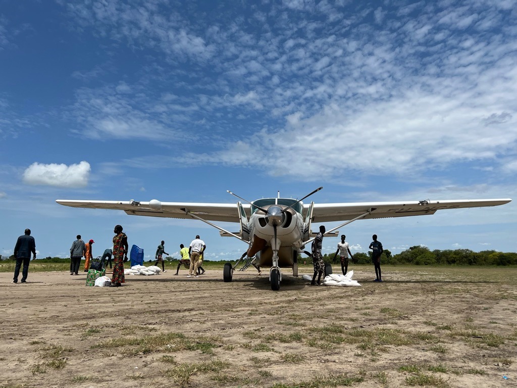 helfende hände - Ein kleines Propellerflugzeug steht auf einer unbefestigten Landebahn unter einem teilweise bewölkten Himmel. In der Nähe stehen mehrere Menschen, von denen einige Taschen und Gegenstände tragen, während andere Vorräte in das Flugzeug ein- oder ausladen. Im Hintergrund sind Büsche und offenes Gelände zu sehen. helfende hände ggmbh hamburg