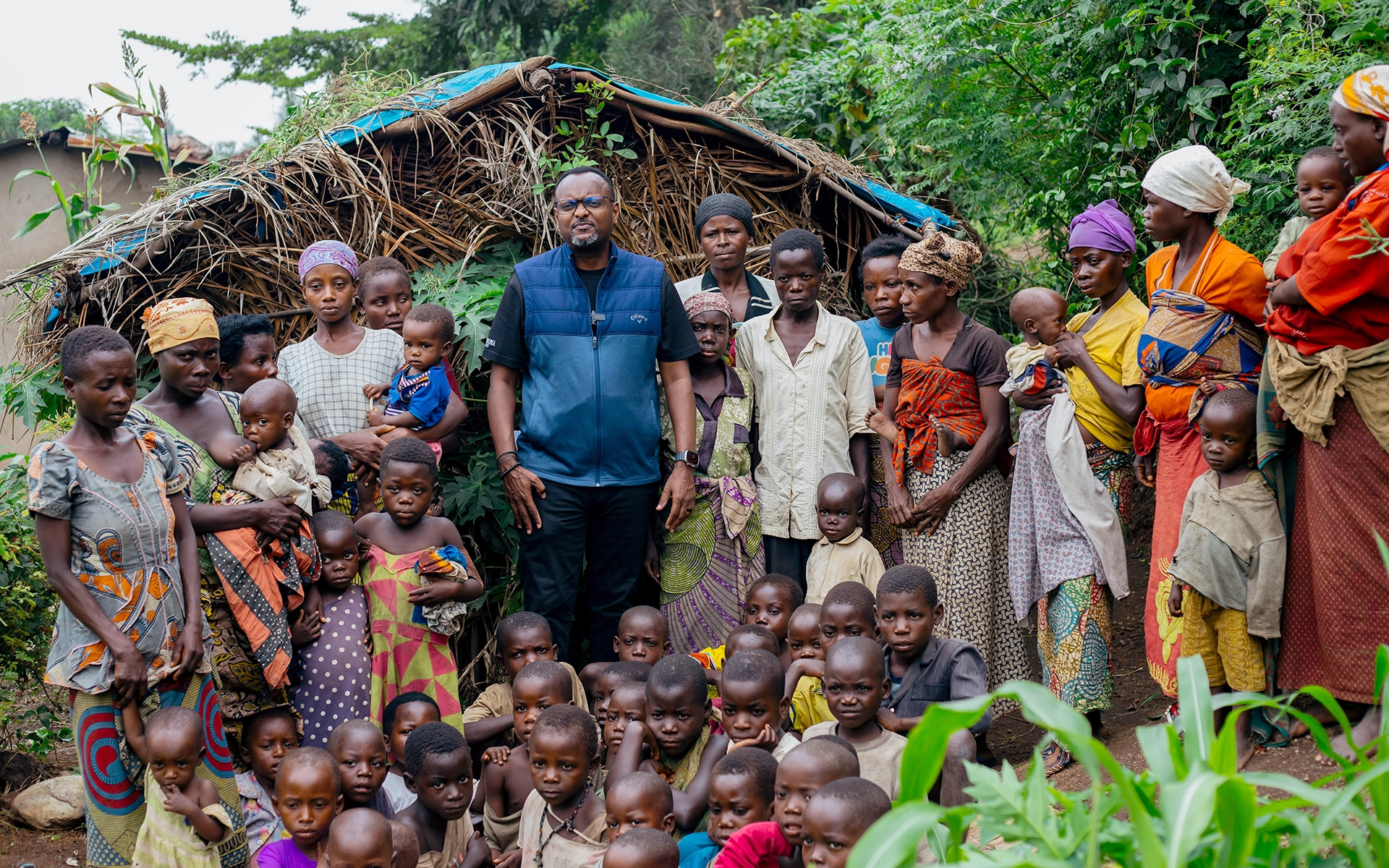 helfende hände - Eine große Gruppe von Erwachsenen und Kindern, darunter auch Claude Nikondea, steht gemeinsam vor einer einfachen, strohgedeckten Hütte, die von Grün umgeben ist. Die meisten blicken in die Kamera, während die Erwachsenen kleine Kinder im Arm halten oder neben ihnen stehen. helfende hände ggmbh hamburg