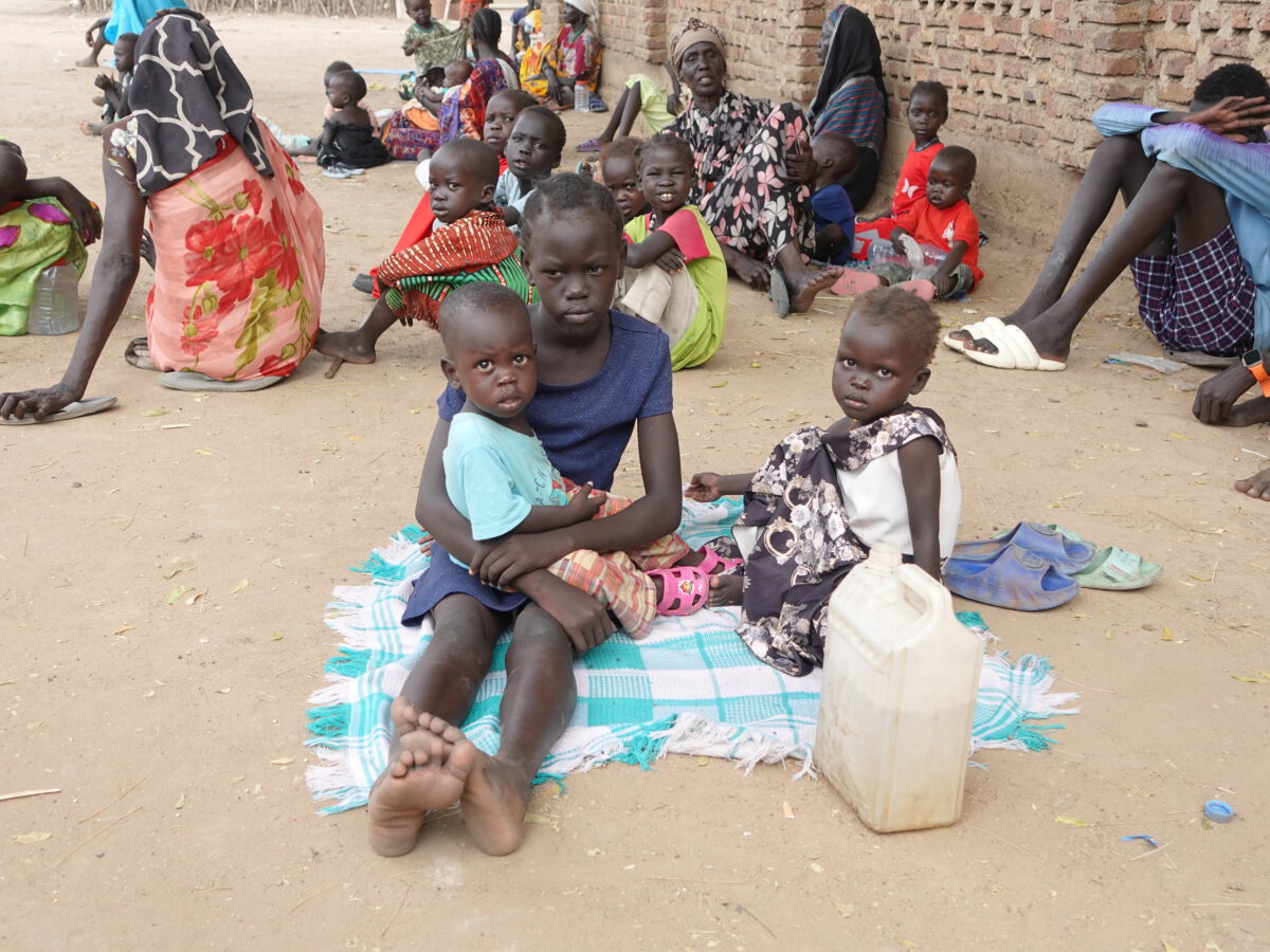 helfende hände - Eine Gruppe von Kindern und Frauen sitzt auf dem Boden in der Nähe einer Backsteinmauer. Ein älteres Kind hält ein jüngeres Kind auf dem Schoß, während ein anderes daneben sitzt. Vor ihnen steht ein Plastikbehälter auf dem Boden. helfende hände ggmbh hamburg