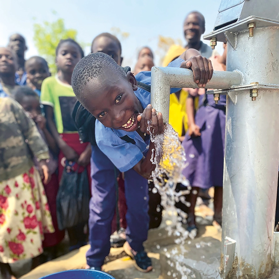 helfende hände - Ein kleiner Junge in einem blauen Hemd trinkt Wasser aus einer Metallhandpumpe und lächelt dabei in die Kamera. Im Hintergrund stehen mehrere Kinder und Erwachsene und schauen zu. Wasser spritzt aus der Pumpe in einen blauen Behälter darunter. helfende hände ggmbh hamburg