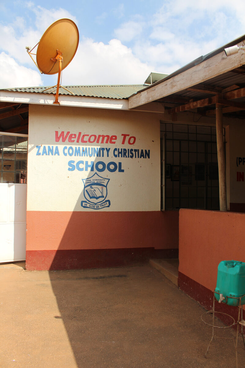 helfende hände - An der Außenwand eines Gebäudes ist ein Schild mit der Aufschrift „Willkommen an der Zana Community Christian School“ zu sehen, unter dem sich ein Logo befindet. Über dem Schild ist eine gelbe Satellitenschüssel angebracht. Im Vordergrund steht eine grüne Handwaschstation. helfende hände ggmbh hamburg