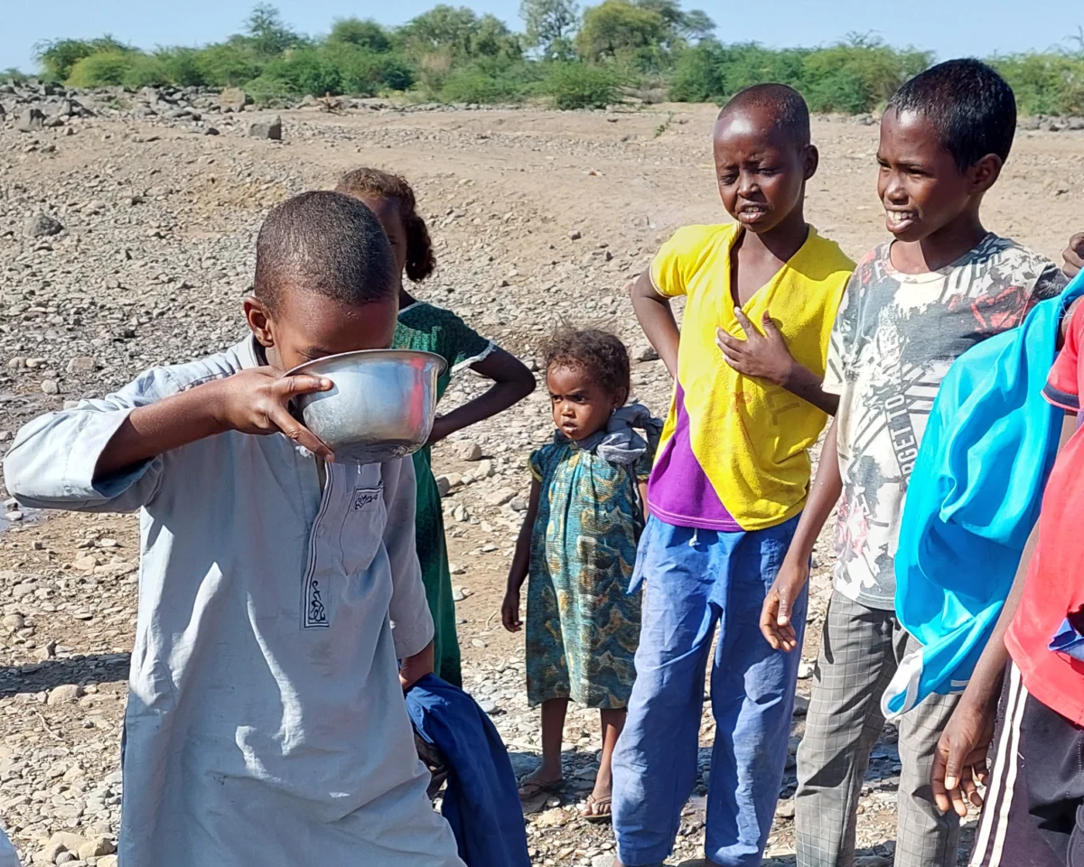 helfende hände - Eine Gruppe von Kindern steht draußen auf trockenem, felsigem Boden. Ein Kind im Vordergrund trinkt aus einer Metallschüssel, während die anderen zusehen. Im Hintergrund sind unter einem klaren Himmel vereinzelte grüne Sträucher zu sehen. helfende hände ggmbh hamburg