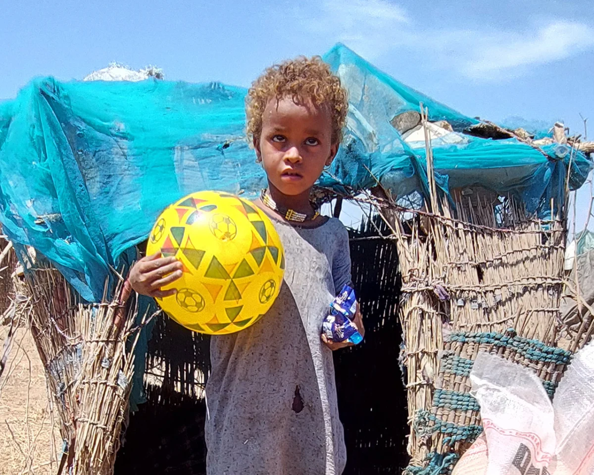 helfende hände - Ein kleines Kind steht vor einer provisorischen Unterkunft aus Stöcken und einer blauen Plane und hält einen gelben Fußball und einen Snack in der Hand. Die Umgebung wirkt trocken und sonnig. helfende hände ggmbh hamburg