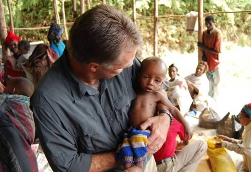 helfende hände - Ein Mann in einem grauen Hemd hält ein unterernährtes Baby im Arm, das in eine gestreifte Decke gewickelt ist. Im Hintergrund sitzen weitere Erwachsene und Kinder und versammeln sich im Freien, wo Holzpfosten und Grünpflanzen zu sehen sind. helfende hände ggmbh hamburg