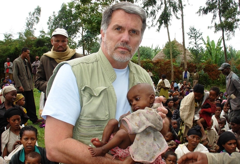 helfende hände - Monte E. Wilson, Einsatzleiter für Nothilfe bei „Helfende Hände International“, hält ein kleines Kind im Arm, während er draußen inmitten einer großen Gruppe sitzender und stehender Menschen steht. Im Hintergrund sind Bäume und rustikale Hütten zu sehen. helfende hände ggmbh hamburg