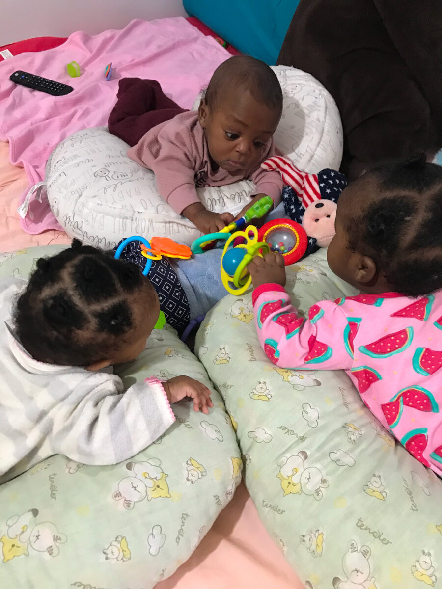 helfende hände - Drei Babys liegen auf Kissen auf einem Bett und sitzen einander gegenüber. Zwei von ihnen spielen mit buntem Spielzeug, während das dritte Baby zusieht. Im Hintergrund sind Stofftiere und eine Fernbedienung zu sehen. helfende hände ggmbh hamburg