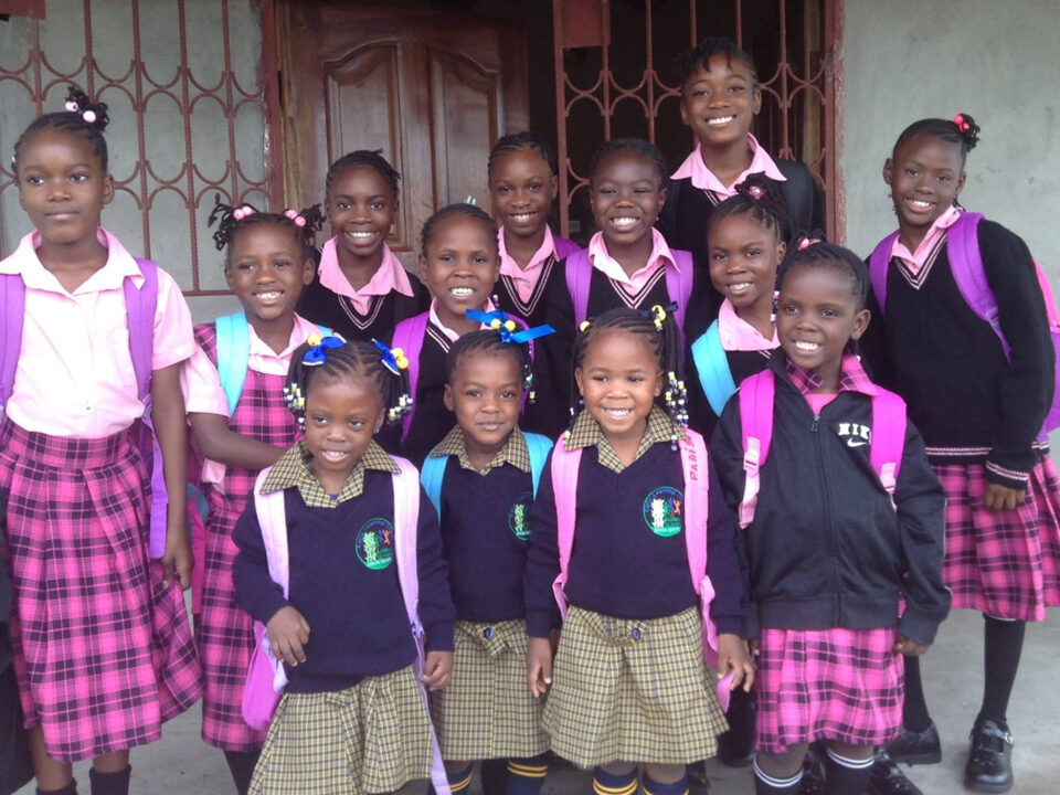 helfende hände - A group of schoolgirls wearing uniforms and backpacks stand in rows, smiling at the camera. The uniforms include plaid skirts and sweaters with school emblems. They are standing outside a building with a barred door. helfende hände ggmbh hamburg