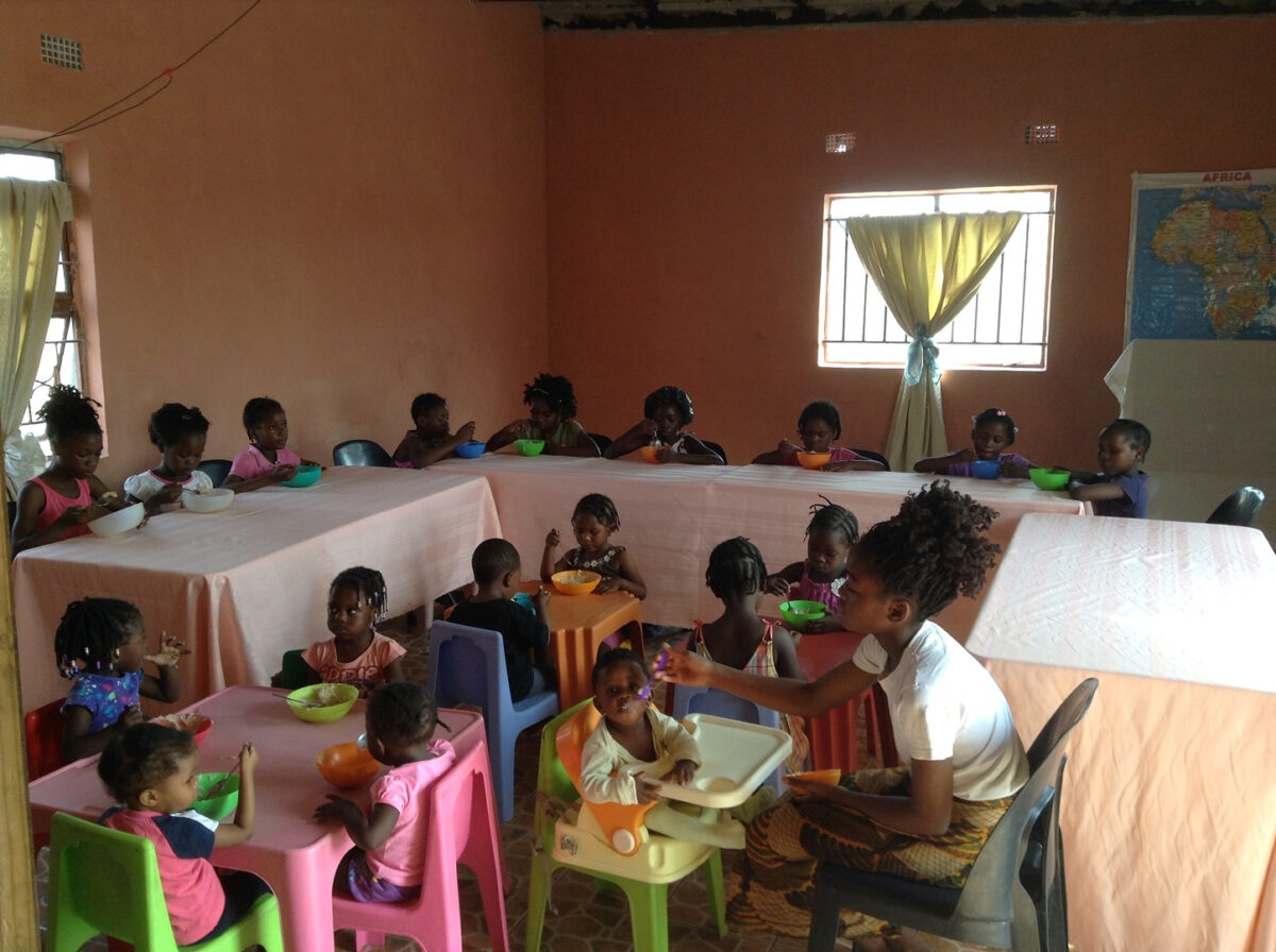 helfende hände - Kinder sitzen in einem Klassenzimmer oder einer Kindertagesstätte an mehreren Tischen und essen aus Schüsseln. Im Vordergrund sitzt ein Erwachsener bei den jüngsten Kindern. Der Raum hat pfirsichfarbene Wände und an der Wand hängt eine Weltkarte. helfende hände ggmbh hamburg