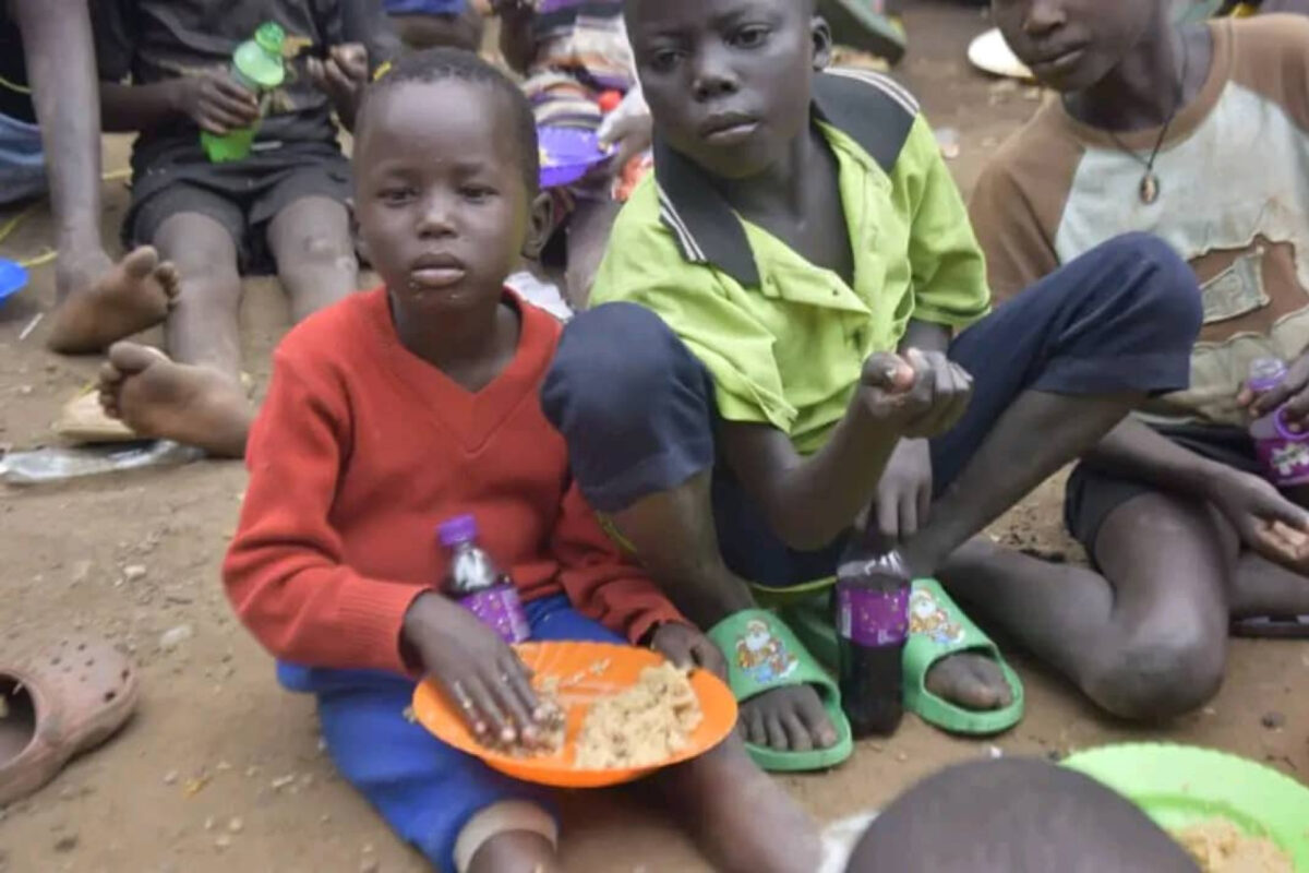 helfende hände - Eine Gruppe von Kindern sitzt draußen auf dem Boden. Ein Kind in einem roten Pullover und einer blauen Hose hält einen orangefarbenen Teller mit Essen und eine lila Trinkflasche in der Hand, während ein anderes Kind in einem grünen T-Shirt in Richtung Kamera winkt. helfende hände ggmbh hamburg