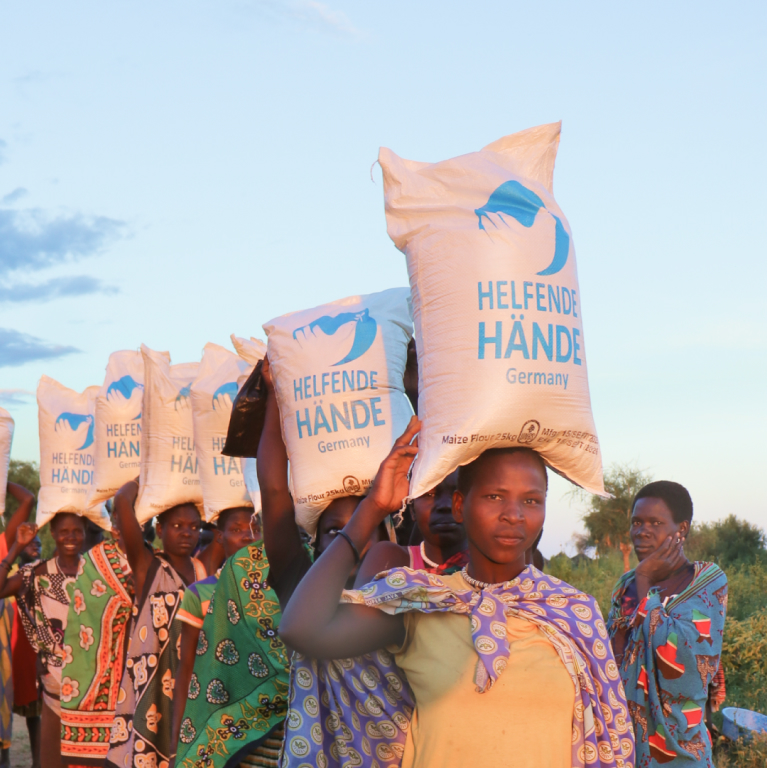helfende hände - Eine Gruppe von Menschen steht im Freien, jeder trägt einen großen weißen Sack mit der Aufschrift „Helfende Hände Germany“ auf dem Kopf. Die Menschen sind in bunte Kleidung gekleidet, und der Himmel ist klar. helfende hände ggmbh hamburg