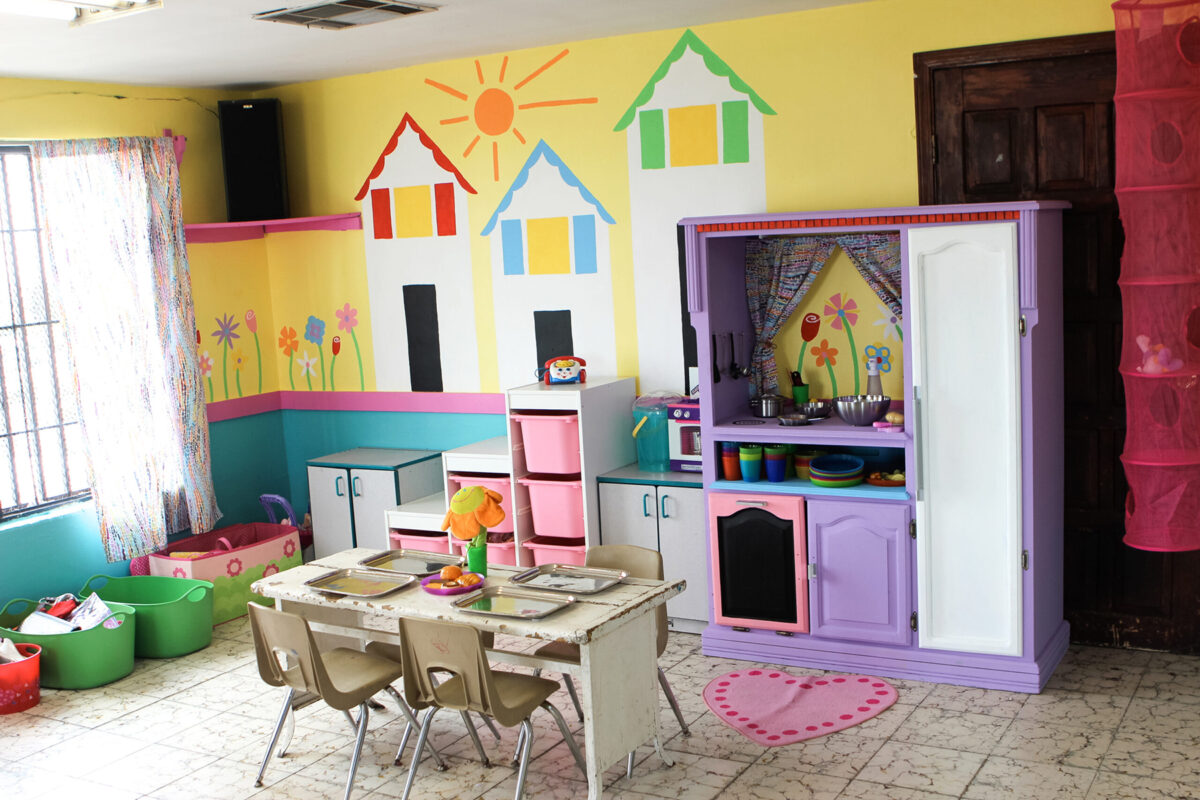 helfende hände - Ein farbenfrohes Spielzimmer mit kleinen Tischen und Stühlen, Spielzeugküchen, Aufbewahrungskörben und bunten Wandbildern mit Häusern, Blumen und einer Sonne. Der Raum wird durch ein Fenster mit transparenten Vorhängen hell beleuchtet. helfende hände ggmbh hamburg