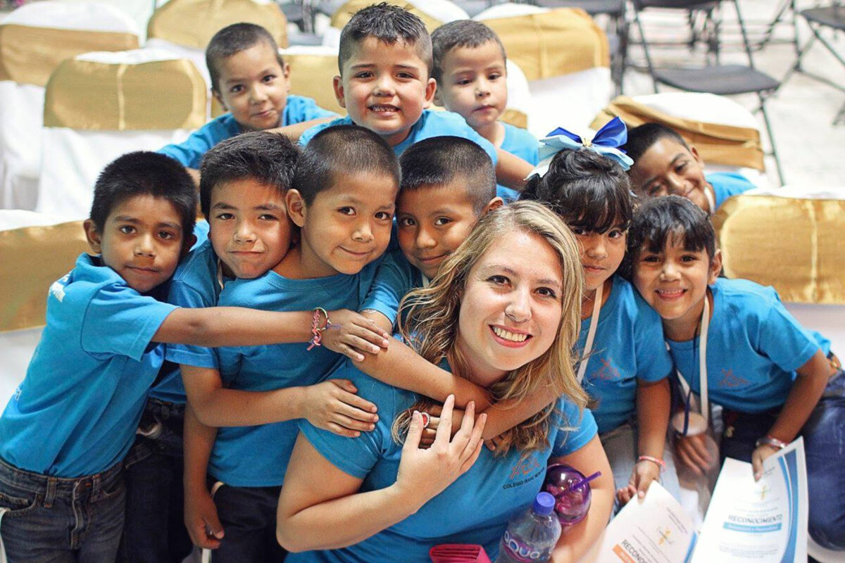 helfende hände - Eine Gruppe kleiner Kinder, die alle einheitliche blaue T-Shirts tragen, versammelt sich um eine lächelnde Frau, die vorne sitzt. Die Kinder umarmen sie und lächeln in die Kamera. Sie scheinen sich in einem hell beleuchteten Innenraum zu befinden. helfende hände ggmbh hamburg