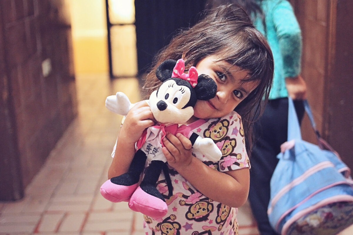 helfende hände - Ein junges Mädchen in einem Pyjama mit Affenmuster steht drinnen und hält ein Minnie-Maus-Plüschtier dicht an ihr Gesicht. Im Hintergrund ist teilweise ein weiteres Kind mit einer blauen Tasche zu sehen. helfende hände ggmbh hamburg