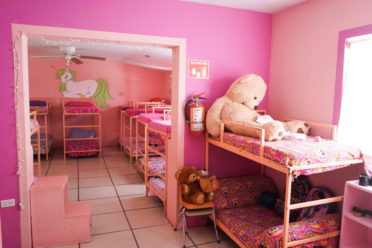helfende hände - Ein hell erleuchteter rosa Raum mit mehreren Etagenbetten, auf einigen liegen Teddybären. An einer Wand ist ein Wandbild mit einem Einhorn zu sehen. Dort steht ein Stuhl mit Stofftieren, ein Feuerlöscher und ein Fenster, durch das Tageslicht hereinfällt. helfende hände ggmbh hamburg