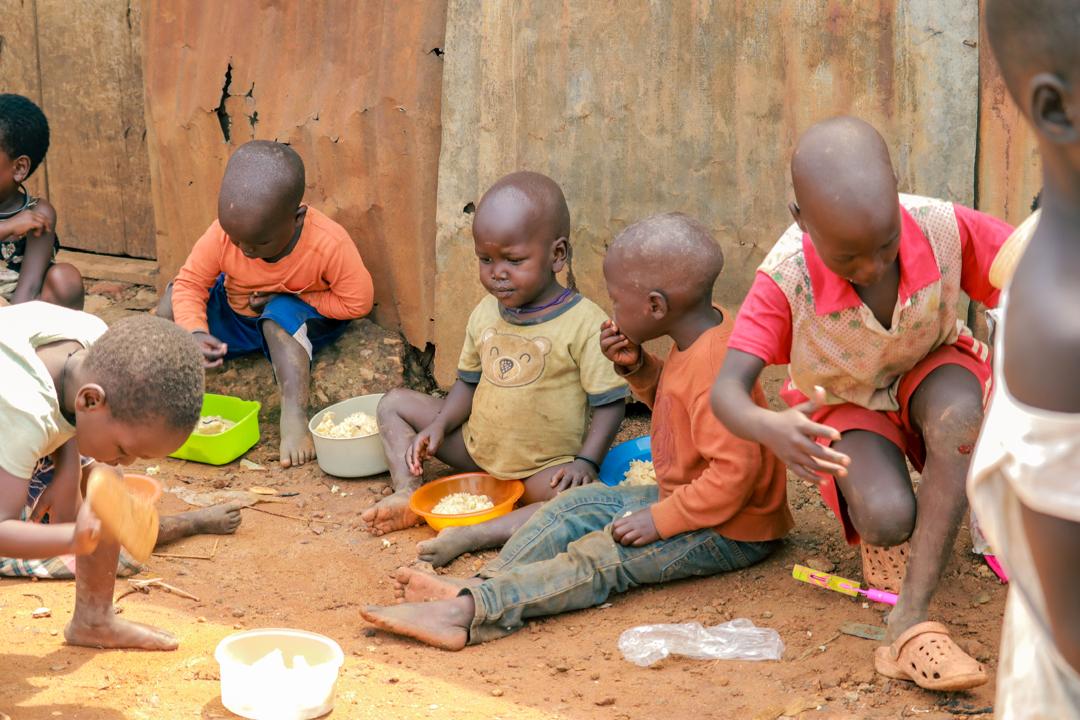 helfende hände - Eine Gruppe kleiner Kinder sitzt draußen auf dem Boden und isst aus Schüsseln. Sie sind barfuß und tragen Freizeitkleidung; im Hintergrund ist eine verrostete Metallwand zu sehen. Einige Kinder sind ganz auf ihr Essen konzentriert. helfende hände ggmbh hamburg