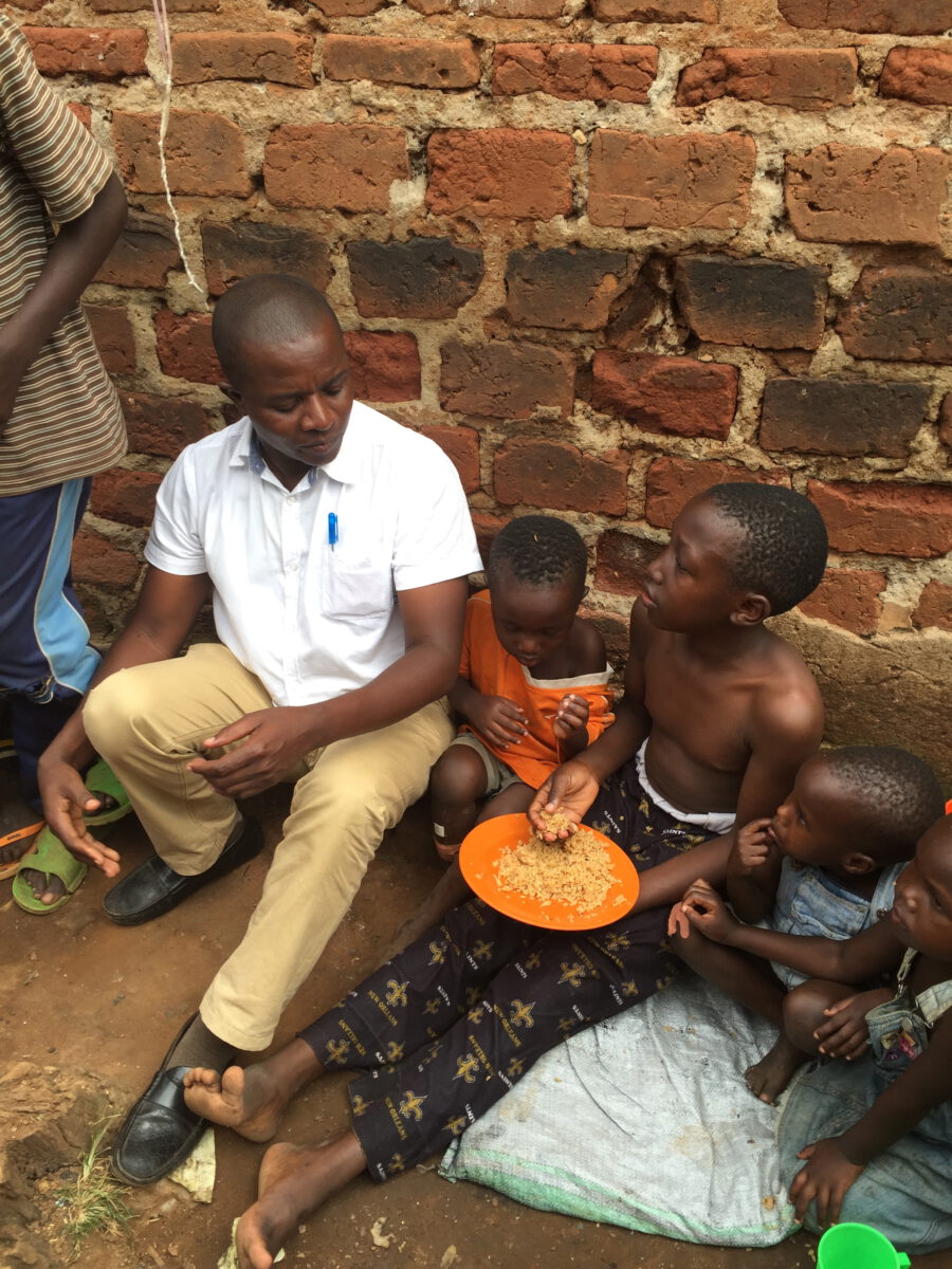 helfende hände - Ein Mann in einem weißen Hemd sitzt auf dem Boden neben mehreren Kindern, die vor einer Backsteinmauer von einem orangefarbenen Teller essen. Ein Kind sitzt barfuß mit einer Schüssel da, und die anderen haben sich dicht um es herum versammelt. helfende hände ggmbh hamburg