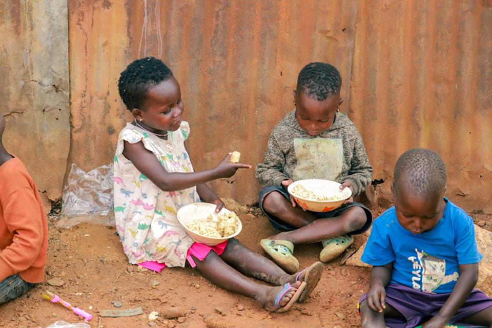 helfende hände - Drei kleine Kinder sitzen auf dem Boden vor einer verrosteten Metallwand und essen aus Schüsseln. Das Kind links trägt ein Kleid und bietet einem anderen Kind etwas zu essen an. Der Boden besteht aus Erde, und in der Nähe liegen einige kleine Gegenstände. helfende hände ggmbh hamburg
