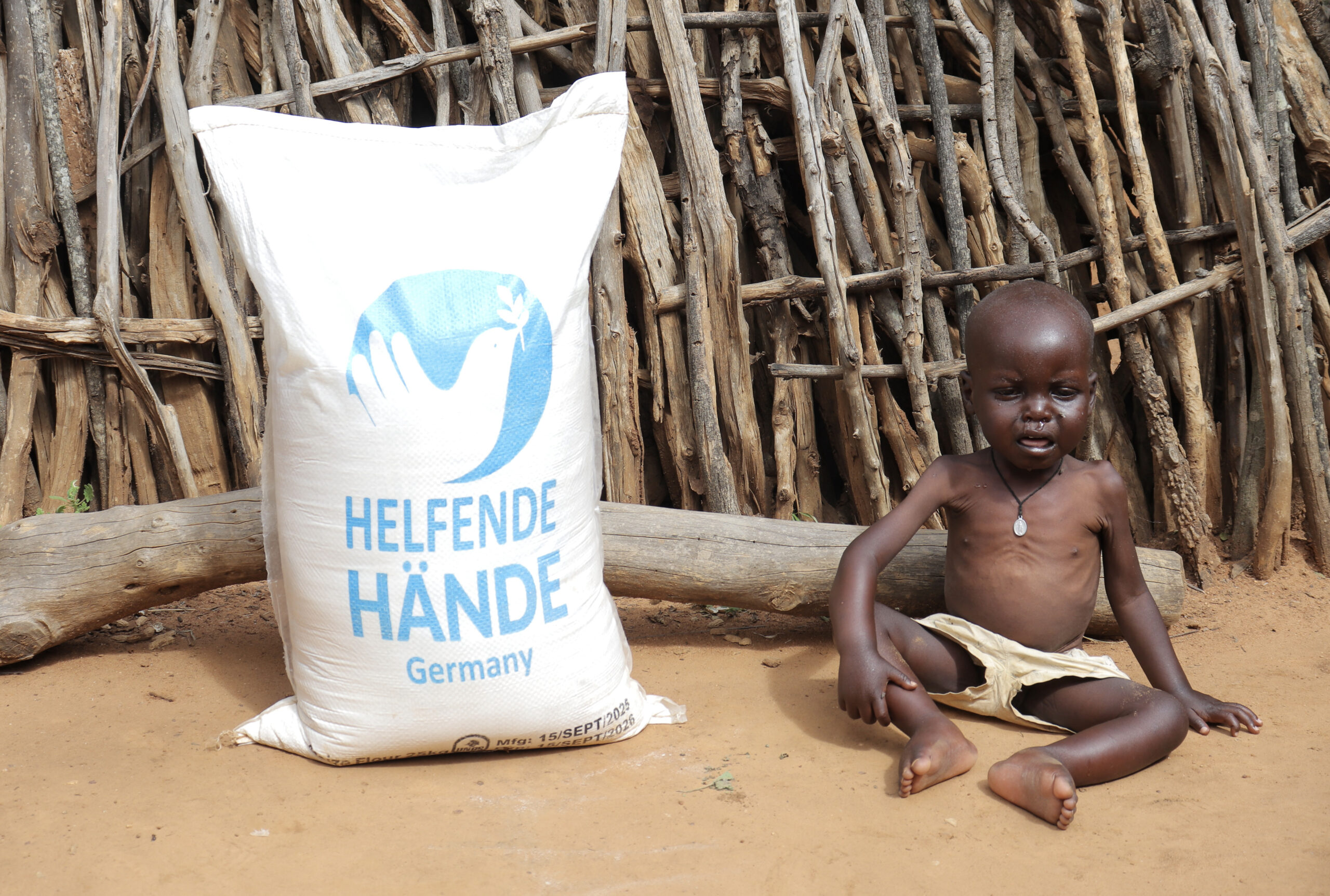 helfende hände - Ein kleines Kind sitzt auf dem Boden neben einem großen weißen Hilfsgutsack mit der Aufschrift „Helfende Hände Germany“ vor einem Holzzaun. Das Kind wirkt dünn und trägt nur Shorts. helfende hände ggmbh hamburg