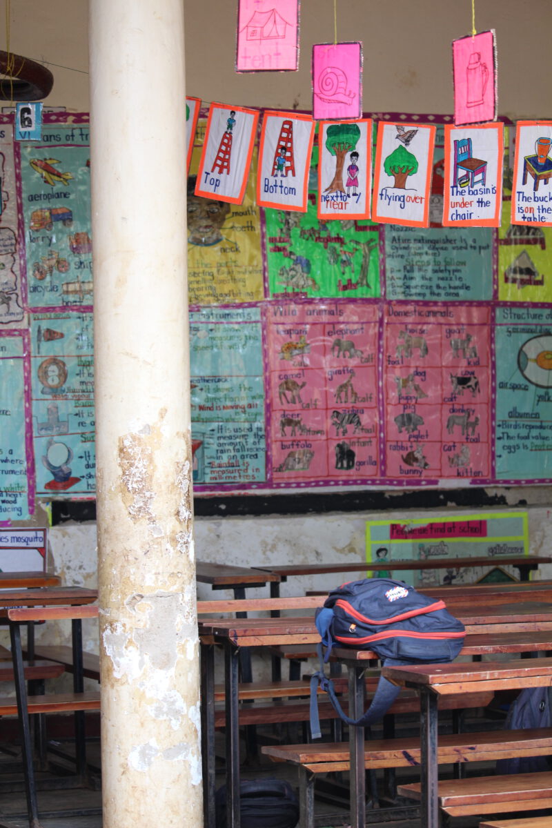 helfende hände - Ein Klassenzimmer mit Holztischen und -bänken, ein blauer Rucksack auf einem Tisch, bunte Lehrplakate und Zeichnungen an der Wand sowie eine abgenutzte Säule im Vordergrund. helfende hände ggmbh hamburg