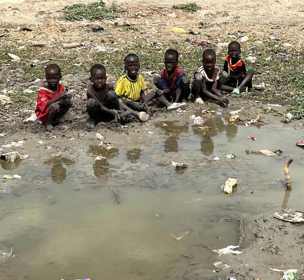helfende hände - Sechs kleine Kinder sitzen auf schlammigem Boden neben einer Pfütze mit schmutzigem Wasser, umgeben von Müll und Unrat. Der Hintergrund ist trocken und karg, mit vereinzelten Grashalmen und spärlicher Vegetation. helfende hände ggmbh hamburg