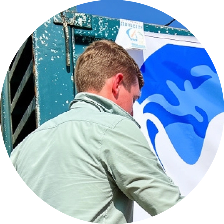 helfende hände - Eine Person mit kurzen, hellbraunen Haaren, die ein hellgrünes Hemd mit Kragen trägt, steht vor einem blau-weißen Plakat auf einer Metallfläche mit Lüftungsschlitzen. Von der Person sind der Rücken und die Seite zu sehen. helfende hände ggmbh hamburg
