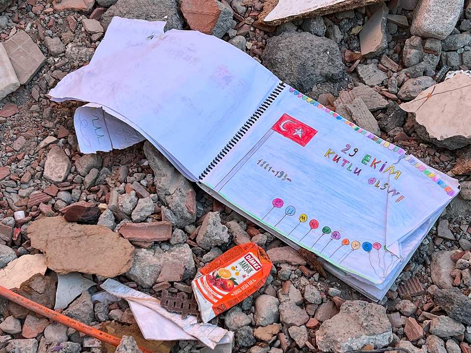 helfende hände - Ein spiralgebundenes Notizbuch mit einer bunten Zeichnung der türkischen Flagge und der Aufschrift „29 Ekim Kutlu Olsun“ liegt offen auf felsigem Boden inmitten von Trümmern, neben einem weggeworfenen Saftkarton. helfende hände ggmbh hamburg