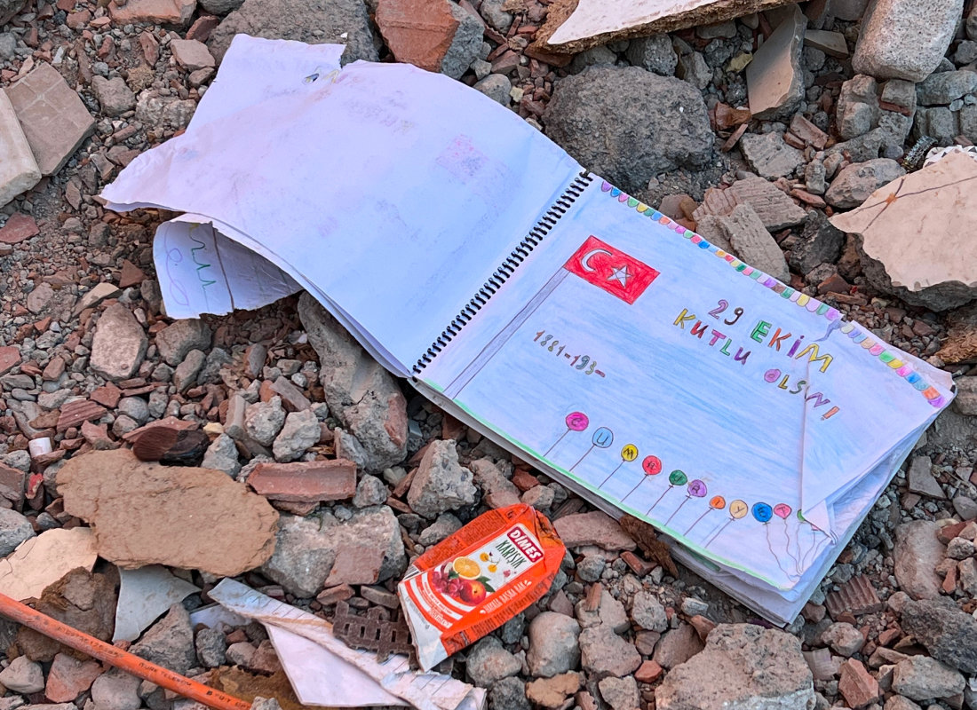 helfende hände - Ein Kinderheft mit einer türkischen Flagge und bunten Zeichnungen liegt offen auf den Trümmern neben einem kleinen Saftpäckchen. Auf der Seite des Hefts steht „29 Ekim Kutlu Olsun“; es scheint sich um ein Schulprojekt zu handeln. helfende hände ggmbh hamburg
