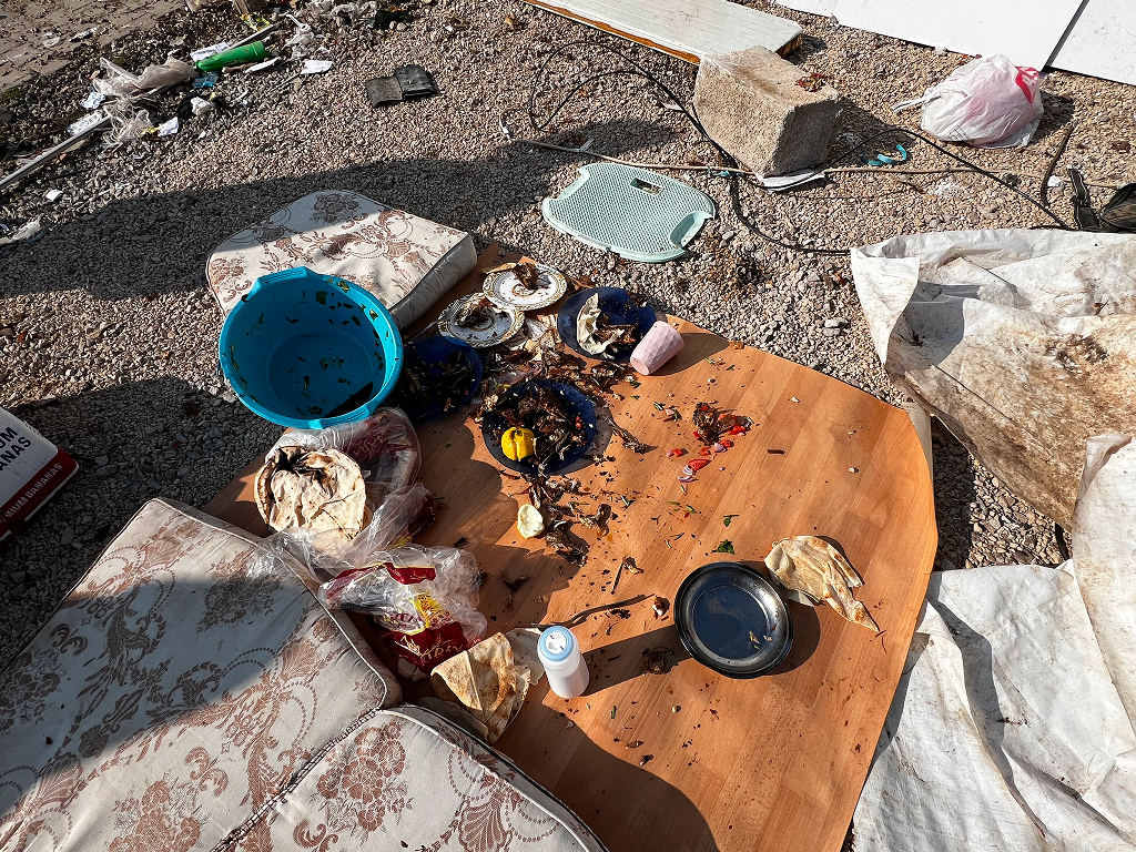 helfende hände - Ein runder Holztisch mit schmutzigem Geschirr, verschütteten Lebensmitteln, Plastiktüten und einer blauen Schüssel steht draußen auf Kies, umgeben von Kissen, Müll und allerlei Unrat. helfende hände ggmbh hamburg