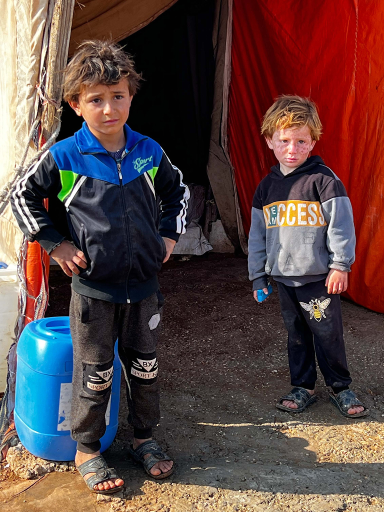 helfende hände - Zwei kleine Kinder stehen vor einem Zelt auf einem unbefestigten Boden, eines mit der Hand in der Hüfte; beide tragen dunkle Trainingsanzüge und Sandalen. Neben ihnen steht ein blauer Wasserbehälter, und hinter ihnen ist das Zelt offen. helfende hände ggmbh hamburg