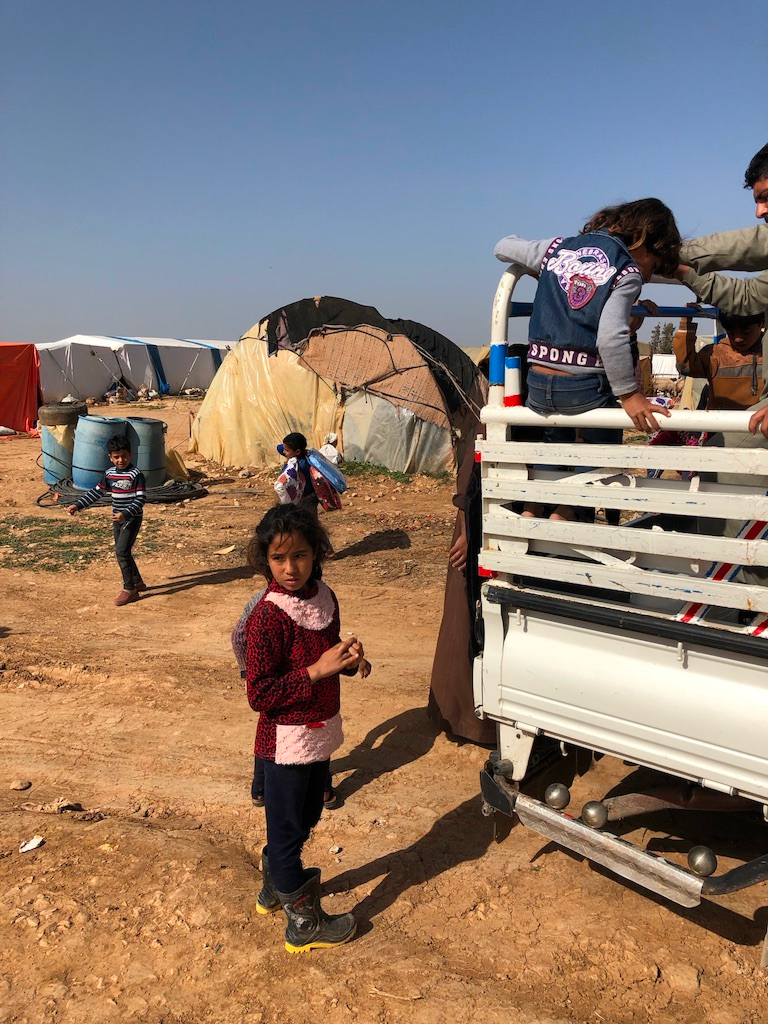 helfende hände - Ein junges Mädchen steht neben einem weißen Lastwagen in einer trockenen, staubigen Gegend, im Hintergrund sind mehrere Kinder und Zelte zu sehen. Andere Kinder klettern auf den Lastwagen. Über ihnen erstreckt sich der blaue Himmel, neben den Zelten stehen Fässer und Behälter. helfende hände ggmbh hamburg