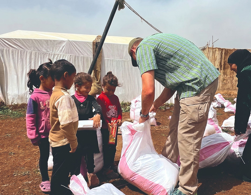 helfende hände - Ein Mann verteilt große weiße Tüten an eine Gruppe von Kindern, die draußen in der Nähe eines weißen Zeltes in einer Reihe stehen. Die Kinder halten einige Gegenstände in den Händen und tragen Freizeitkleidung. Ein weiterer Erwachsener steht in der Nähe. helfende hände ggmbh hamburg