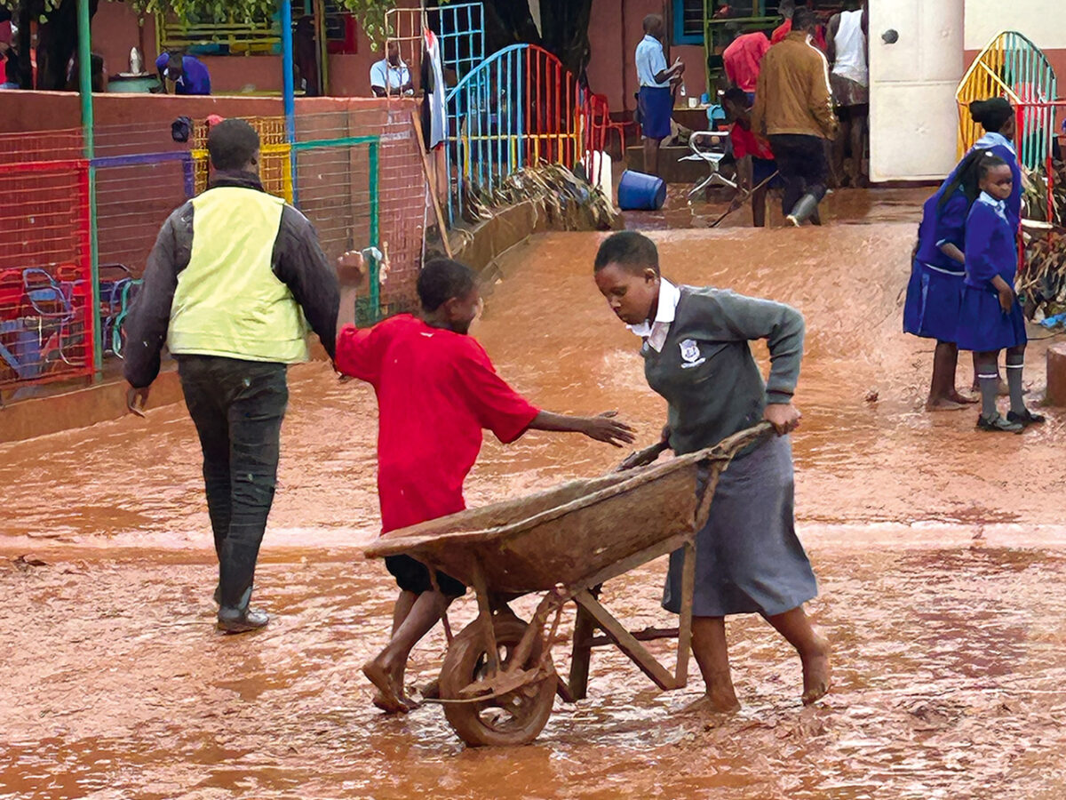 helfende hände - Ein Mädchen in einer grauen Schuluniform schiebt eine hölzerne Schubkarre durch schlammiges Wasser, während ein Junge in einem roten Hemd die Hand nach ihr ausstreckt. In der Nähe gehen weitere Menschen durch das schlammige Gelände, im Hintergrund sind bunte Zäune und Gebäude zu sehen. helfende hände ggmbh hamburg