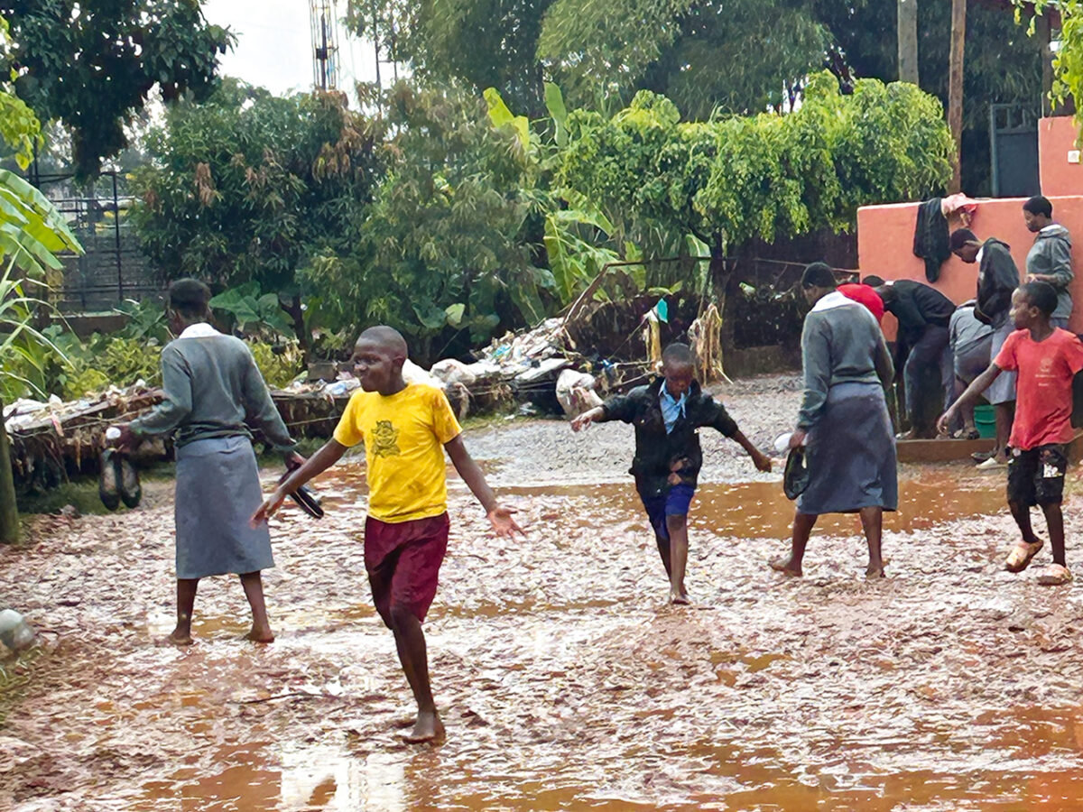 helfende hände - Nach einem Regenschauer laufen und spielen mehrere Kinder und Erwachsene im Freien auf einem schlammigen, durchnässten Gelände, das von Bäumen und Pflanzen umgeben ist. Einige Menschen stehen in der Nähe einer orangefarbenen Mauer, und der Boden ist mit Pfützen und Schlamm bedeckt. helfende hände ggmbh hamburg