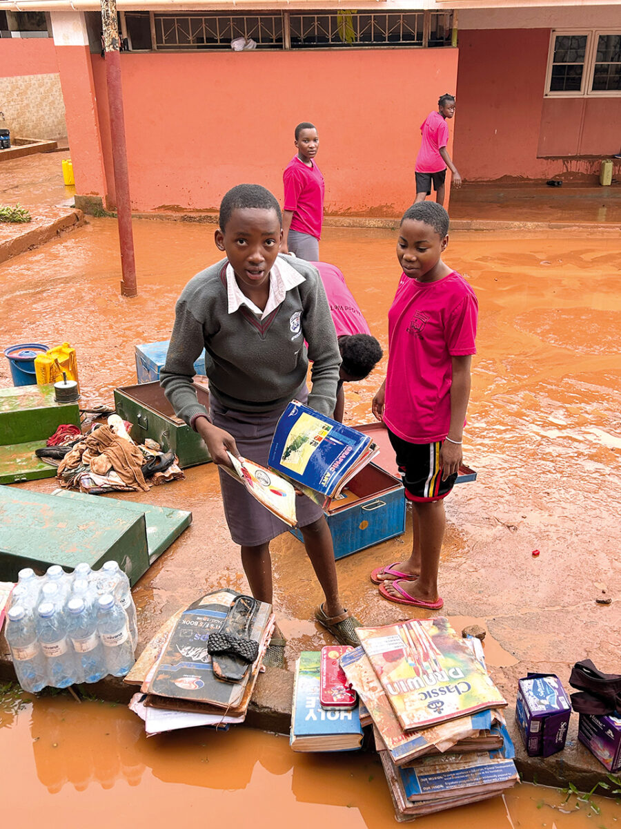helfende hände - Ein Schüler in Schuluniform steht auf einem schlammigen, überfluteten Schulhof und hält nasse Schulbücher in den Händen. Auf dem Boden liegen Stapel beschädigter Bücher, Wasserflaschen und andere Hilfsgüter. Drei weitere Schüler in rosa Hemden stehen in der Nähe. helfende hände ggmbh hamburg