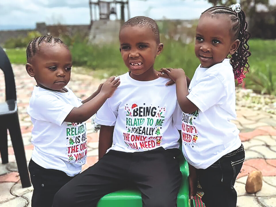 helfende hände - Drei kleine Kinder, die alle einheitliche weiße T-Shirts tragen, stehen und sitzen gemeinsam im Freien auf einer gepflasterten Fläche. Zwei Mädchen stehen zu beiden Seiten eines sitzenden Jungen und haben ihre Hände auf seine Schultern gelegt. helfende hände ggmbh hamburg